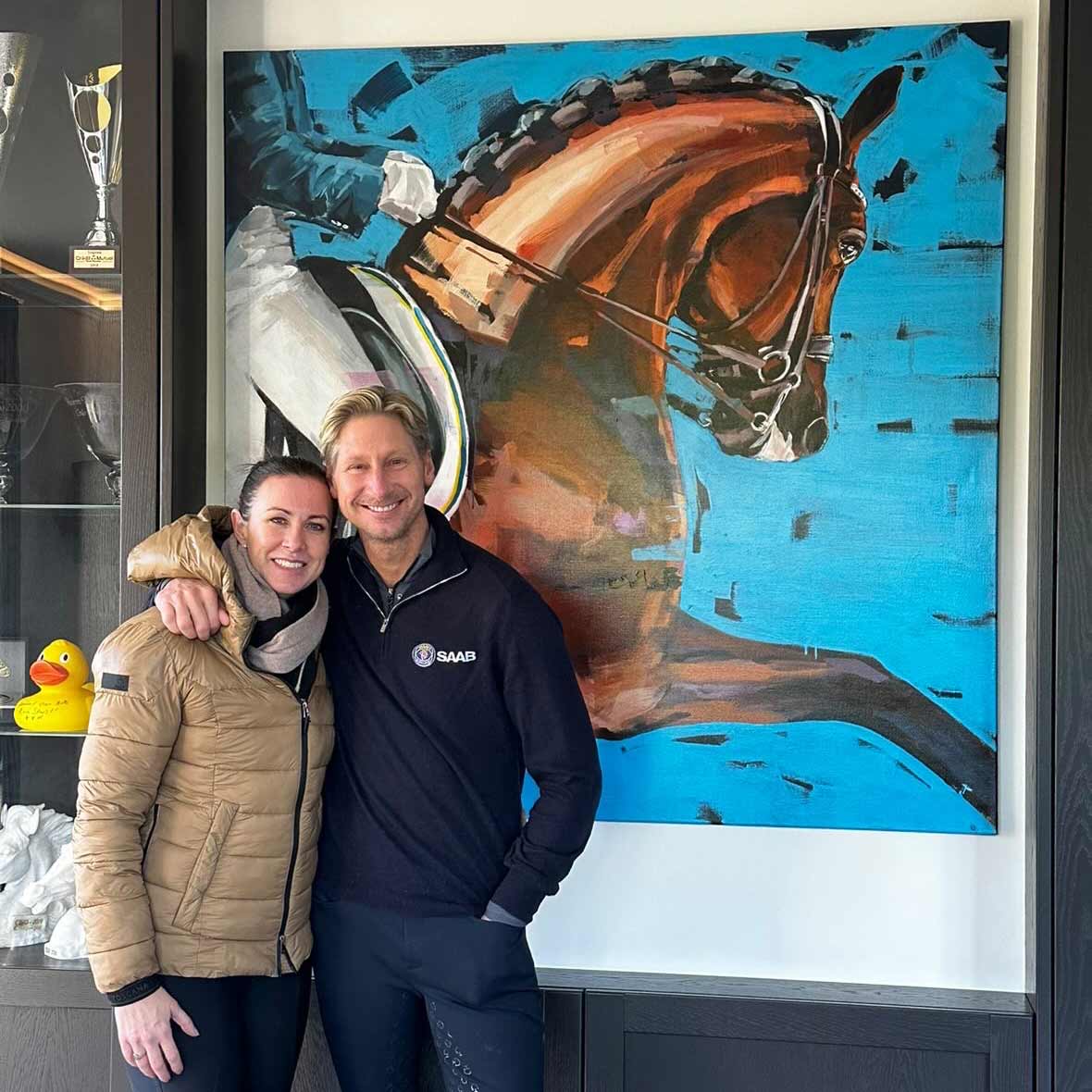 Foto der Weltklasse-Dressurreiter Patrik Kittel und Lyndal Oatley, die lächelnd vor ihrem großformatigen Pferdeporträt des Hengstes „Touchdown“ von Hartmut Hellner stehen; moderne Tiermalerei auf leuchtend blauem Hintergrund in einem privaten, exklusiven Ambiente.