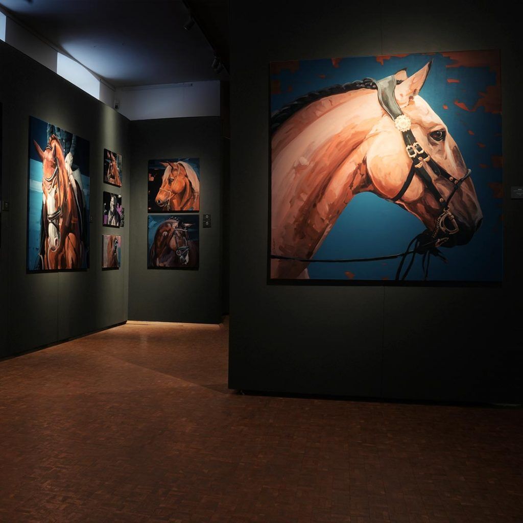Exhibition view from the German Horse Museum with a large-scale horse portrait in the foreground, modern animal painting by Hartmut Hellner on a dark gallery wall, highlighting precise lighting and the expressive profile of the horse.