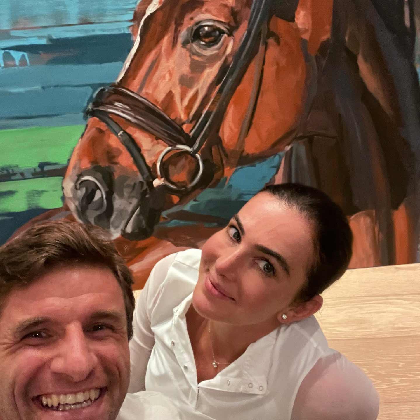Ein Selfie von Thomas Müller und seiner Frau Lisa vor dem großformatigen Pferdeporträt ihres Hengstes D'avie, moderne Pferdemalerei von Hartmut Hellner in warmen Brauntönen, prominent platziert im privaten Wohnzimmer.