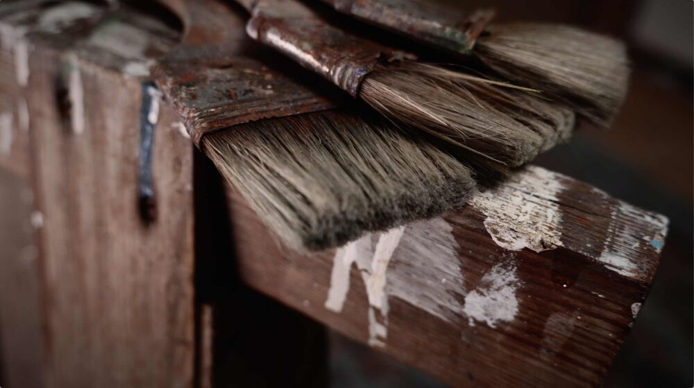 Nahaufnahme von drei breiten, farbbespritzten Pinseln auf einem rustikalen Holzbock im Atelier des Pferde- und Tiermalers Hartmut Hellner, stimmungsvolles Licht betont die Textur der Borsten.